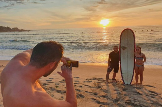 Muž fotí surfaře na pláži při západu slunce.
