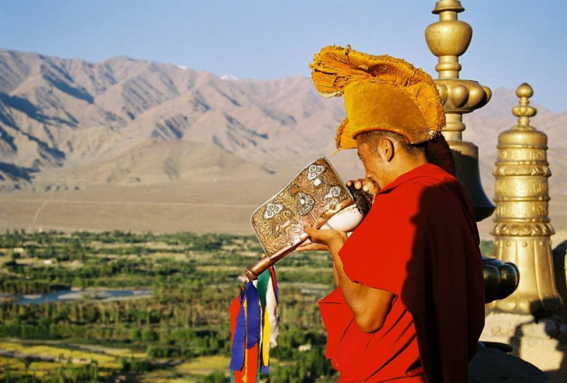 Tibetský mnich v tradičním rouchu troubí v buddhistickém klášteře obklopeném horami. 